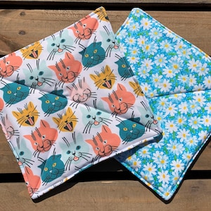 May include: Two square pot holders. One is white with a pattern of colourful cats with their tongues sticking out. The other is blue with a white daisy pattern.