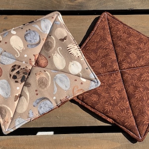 May include: Two brown and beige pot holders. One pot holder has a pattern of cats on a beige background. The other pot holder has a brown and beige leaf pattern.
