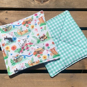 May include: A pair of reversible pot holders. One side features a white background with a colourful pattern of cats, flowers, and butterflies. The other side is a green and white gingham pattern.