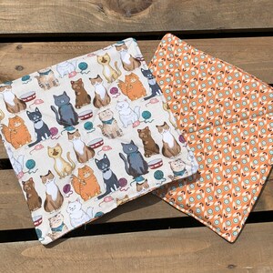 May include: Two fabric squares with a cat theme. One square is white with a pattern of colorful cats, yarn balls, and cat toys. The other square is orange with a repeating geometric pattern.