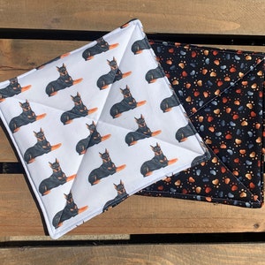 May include: Two white and black patterned pot holders. One pot holder features a repeating pattern of black and tan dogs. The other pot holder features a black background with a repeating pattern of orange and blue paw prints.