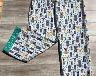 May include: White pajama pants with a pattern of various dog breeds and bone shapes. The pants have a teal blue cuff with a white bone pattern.