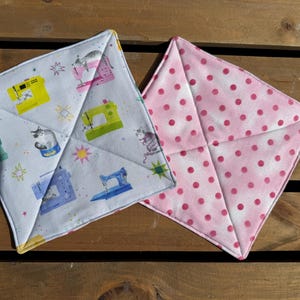May include: Two square fabric bowl cozies. One features a white background with a pattern of colorful sewing machines, cats, and stars. The other is made of pink fabric with a repeating pattern of dark pink polka dots. Both have stitched seams.