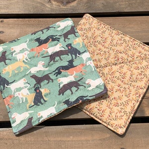 May include: Two green and beige pot holders with a dog pattern. One pot holder has a green background with a pattern of black and brown dogs. The other pot holder has a beige background with a pattern of brown leaves.