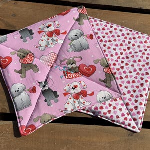 May include: Two pink and white pot holders with a heart pattern and a dog design. The pot holders are made of fabric and have a quilted texture.