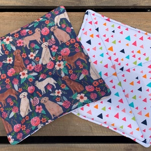 May include: Two fabric pot holders. One is navy blue with a floral pattern and brown and white dogs. The other is white with a pattern of colourful triangles.