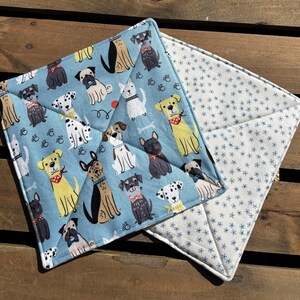 May include: Square quilted potholder with a light blue background featuring various dog breeds in different colors. The reverse side is white with a blue floral pattern. The potholder is designed for kitchen use.