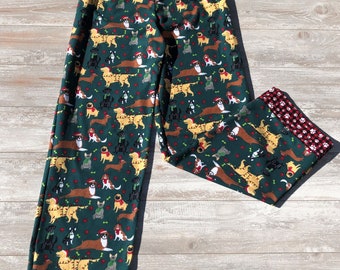 May include: Green pajama pants with a festive pattern of dogs wearing Santa hats and scarves. The pants have red and white plaid cuffs with paw prints.