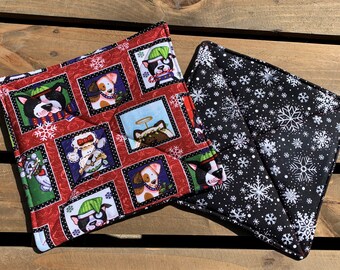 May include: Two square quilted pot holders. One has a red background with framed images of dogs in holiday attire and snowflakes. The other has a black background with white snowflakes. Both have black binding.