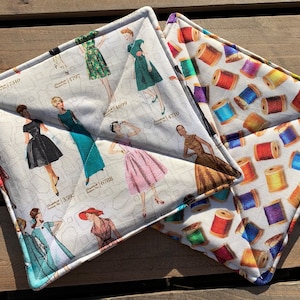 May include: Two white cotton pot holders. One features a pattern of colorful spools of thread. The other features a pattern of vintage fashion illustrations.