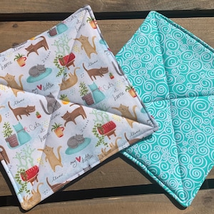 May include: Two square pot holders. One is white with a repeating pattern of cats, plants, and the text "Cats at home". The other is turquoise with a repeating pattern of white swirls.