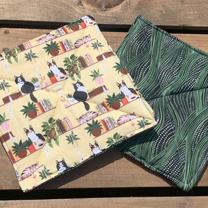 May include: Two pot holders, one with a green wavy pattern and the other with a yellow background and a repeating pattern of cats on bookshelves.