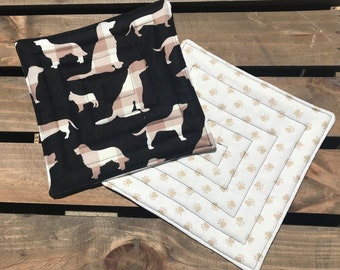 May include: Two quilted pot holders. One is black with a white plaid dog silhouette pattern. The other is white with a tan paw print pattern.