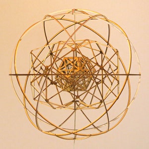 Op de afbeelding: Een gouden, geometrisch, houten sculptuur met een bolvorm. De sculptuur is gemaakt van meerdere elkaar snijdende cirkels en vierkanten.