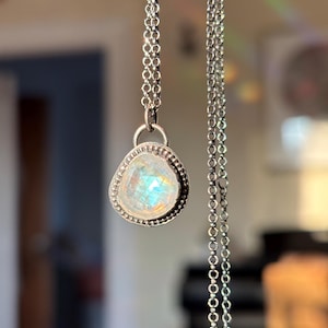May include: A silver necklace with a pendant featuring a pear-shaped, iridescent gemstone. The pendant is set in a textured silver bezel and hangs from a delicate chain. The gemstone displays a range of colors, including blue, yellow, and white.