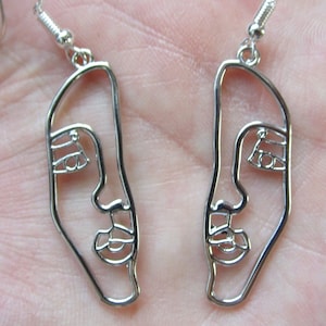 May include: A pair of silver-colored wire earrings. Each earring is a stylized outline of a face, featuring a nose, eye, and mouth. The earrings have a hook closure. The design is minimalist and modern.