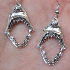 May include: A pair of silver-colored earrings shaped like open shark jaws. Each earring features detailed teeth and a hook for pierced ears. The earrings are designed to dangle and catch the light.