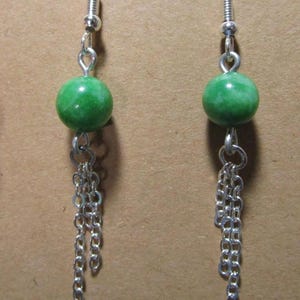 May include: A pair of dangle earrings featuring bright green, round beads and silver-colored chain tassels. The earrings have silver-colored fish hook ear wires. The earrings are displayed against a brown background.