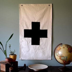 May include: A white canvas flag with a large black plus sign in the center. The flag is hanging on a wall with a globe and plants in the foreground.
