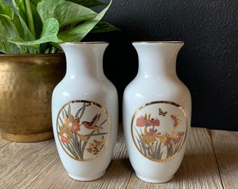 Puede incluir: Dos jarrones de cerámica blanca con ribetes dorados y diseños florales. Un jarrón presenta un pájaro y el otro mariposas.