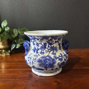 May include: A white and blue ceramic vase with a floral design. The vase has two blue handles and a wide opening. The floral pattern includes large peony-like flowers and smaller leaf and vine details.