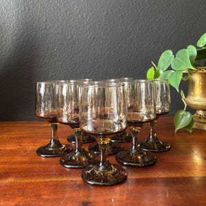 May include: A set of eight vintage-style wine glasses. Each glass features a brown-tinted bowl, a slender stem, and a circular base. The glasses are arranged on a wooden surface, with a dark background and a touch of greenery.
