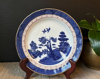 May include: A blue and white porcelain plate with a willow tree design. The plate has a gold rim and a red and gold border.