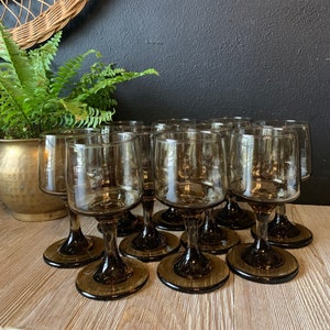 May include: A set of 12 brown glass wine glasses with a clear bowl and a thick stem. The glasses are arranged in a circle on a light brown wooden surface.