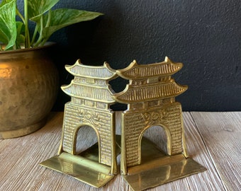 May include: Two brass pagoda-style bookends. Each bookend features a detailed architectural design with a brick facade, arched doorway, and tiered roof. The bookends are placed on a light wood surface, with a plant in a brass pot in the background.