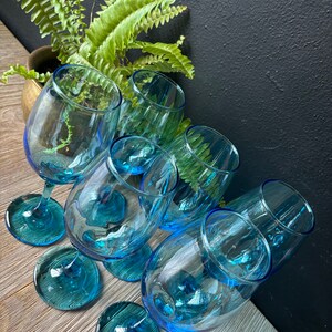 May include: Six blue glass wine glasses with a textured design. The glasses are arranged in a circle on a wooden surface.