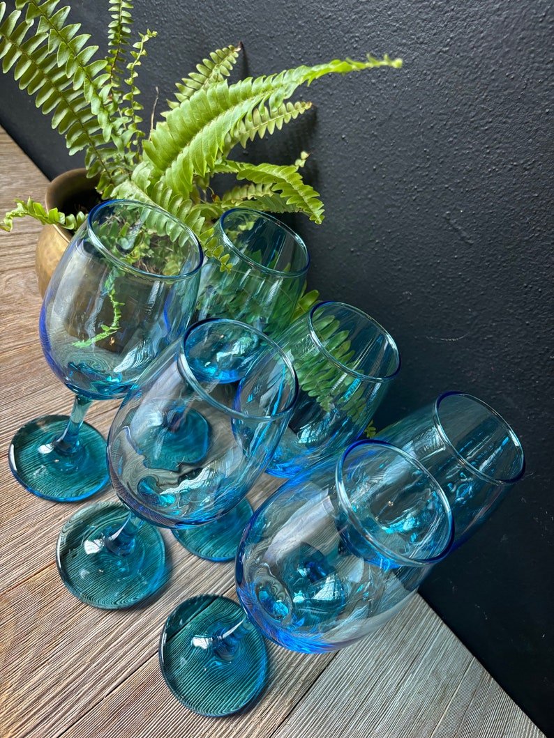 May include: Six blue glass wine glasses with a textured design. The glasses are arranged in a circle on a wooden surface.
