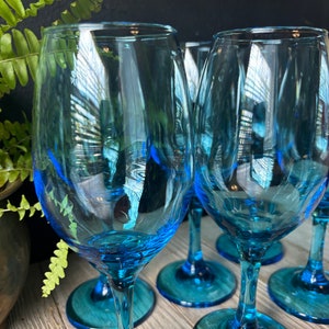 May include: Set of six blue glass wine glasses with a teal colored base. The glasses are clear and have a rounded bowl shape.