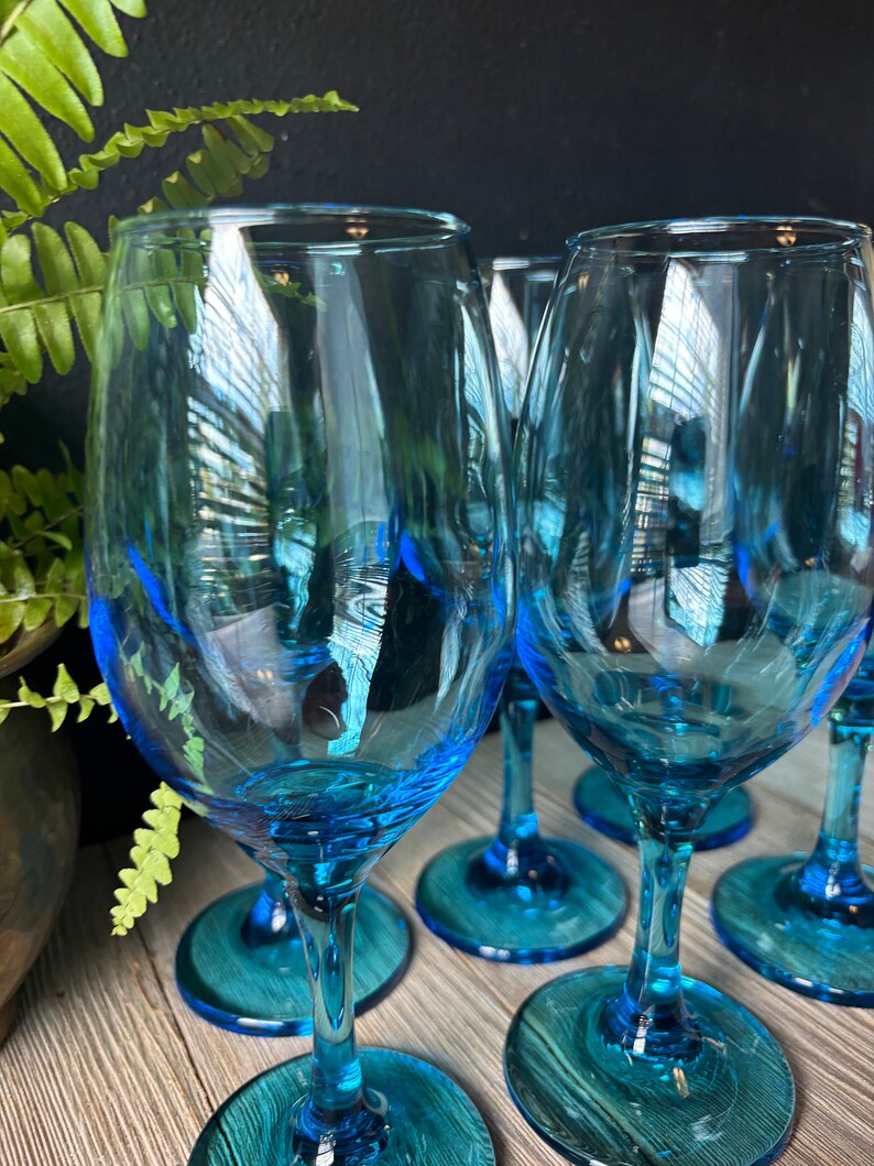 May include: Set of six blue glass wine glasses with a teal colored base. The glasses are clear and have a rounded bowl shape.