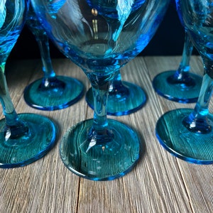 May include: Set of six blue glass wine glasses with a clear base. The glasses are arranged in a semi-circle on a wooden surface.