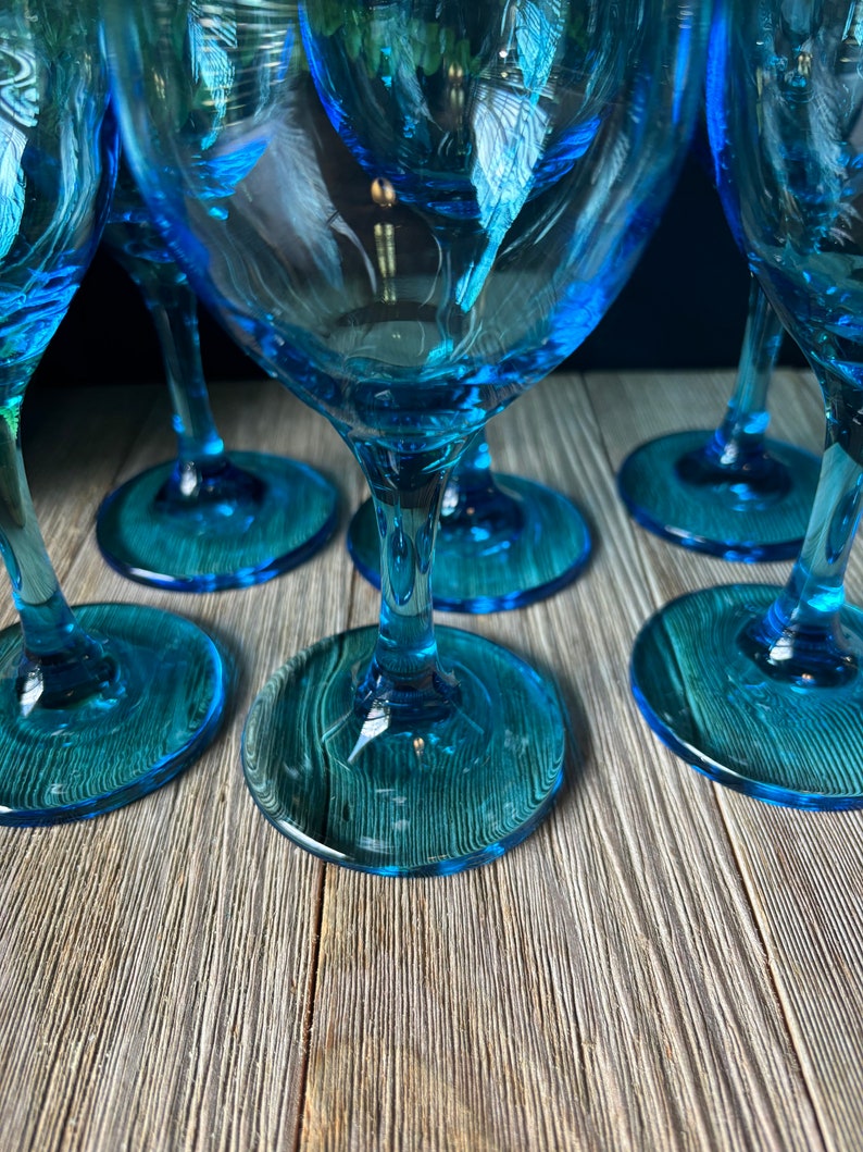 May include: Set of six blue glass wine glasses with a clear base. The glasses are arranged in a semi-circle on a wooden surface.