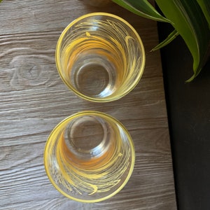May include: Two clear glass drinking glasses with yellow painted designs. The glasses have a rounded bottom and a straight, cylindrical top.