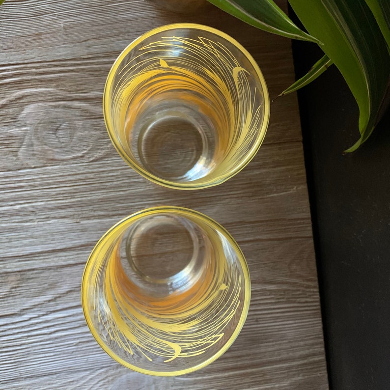 May include: Two clear glass drinking glasses with yellow painted designs. The glasses have a rounded bottom and a straight, cylindrical top.