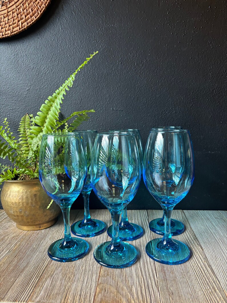 May include: Six turquoise blue wine glasses on a wooden surface. The glasses are clear and have a slight blue tint.
