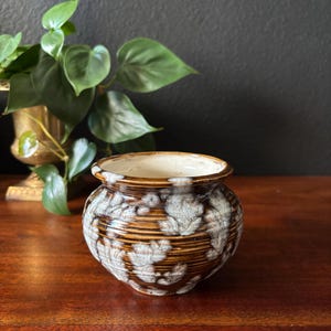 May include: A small, brown ceramic pot with a white and brown floral design. The pot is sitting on a wooden surface.