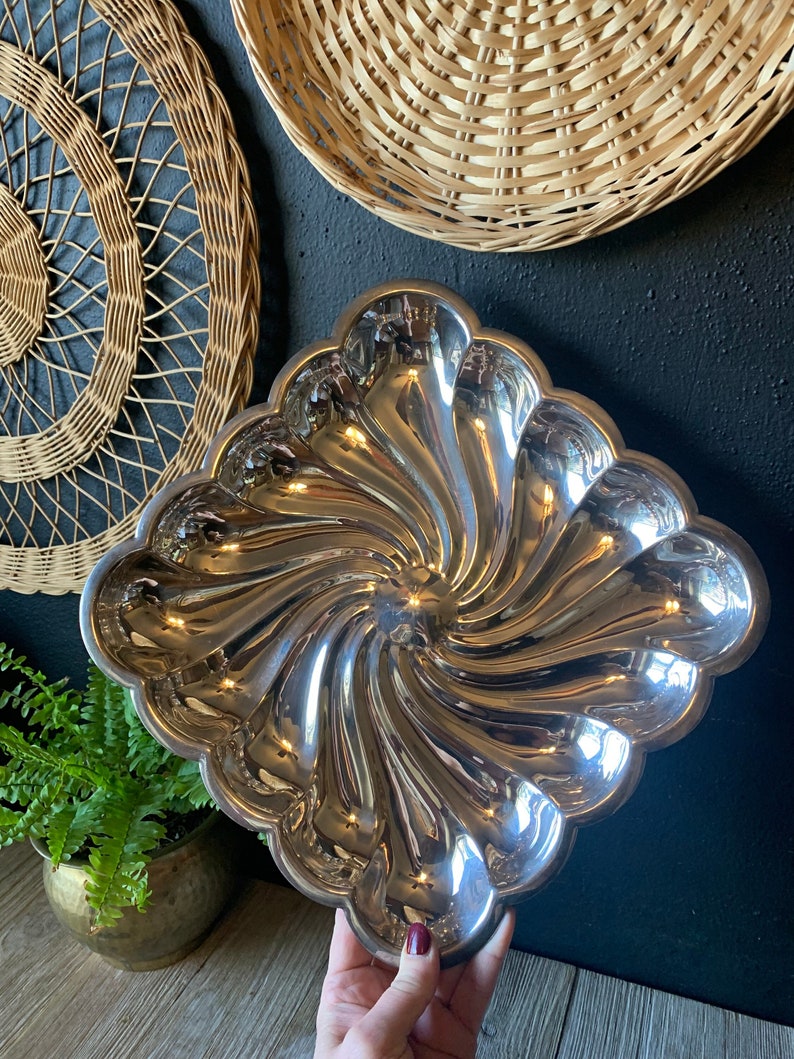 May include: A silver, swirl-shaped serving dish with a scalloped edge. The dish is made of metal and has a shiny, reflective surface.