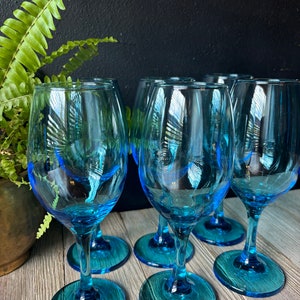 May include: Six blue glass wine glasses with a clear base. The glasses are arranged in a semi-circle on a wooden surface.