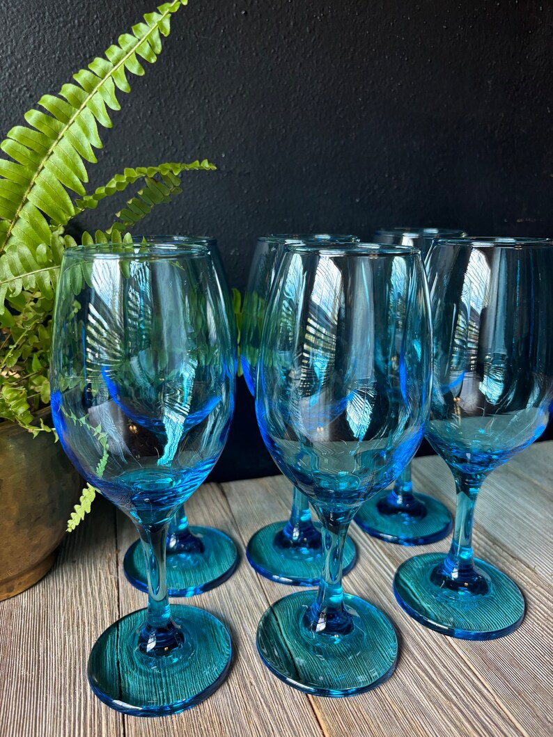 May include: Six blue glass wine glasses with a clear base. The glasses are arranged in a semi-circle on a wooden surface.