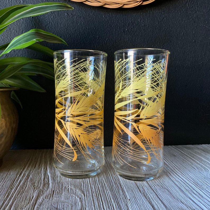 May include: Two clear glass drinking glasses with a gold wheat design. The glasses are on a wooden surface.