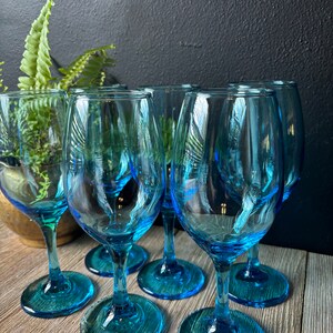 May include: Six blue glass wine glasses with a teal tint. The glasses are arranged in a semi-circle on a wooden surface.