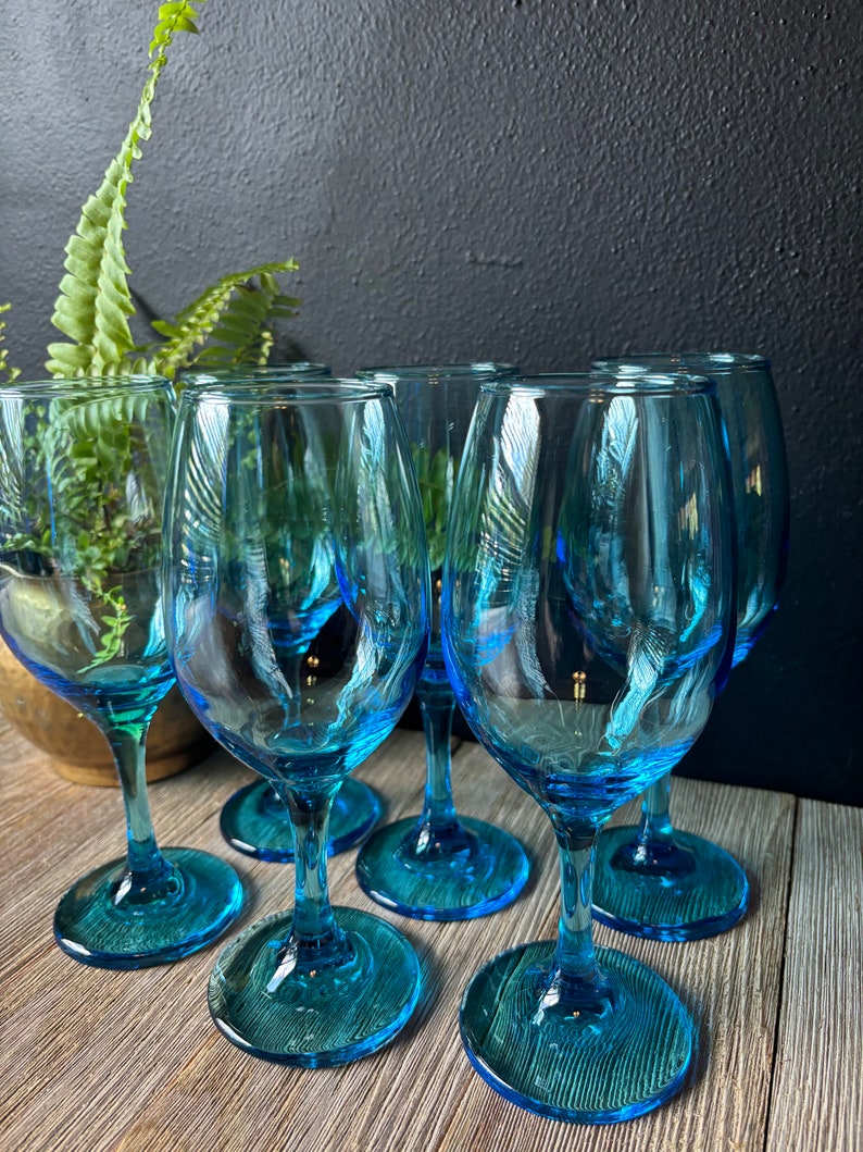 May include: Six blue glass wine glasses with a teal tint. The glasses are arranged in a semi-circle on a wooden surface.