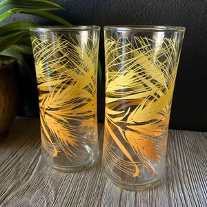 May include: Two clear glass tumblers with a yellow wheat design. The glasses are empty and sitting on a wooden surface.