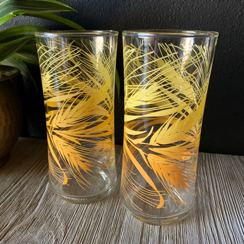 May include: Two clear glass tumblers with a yellow wheat design. The glasses are empty and sitting on a wooden surface.