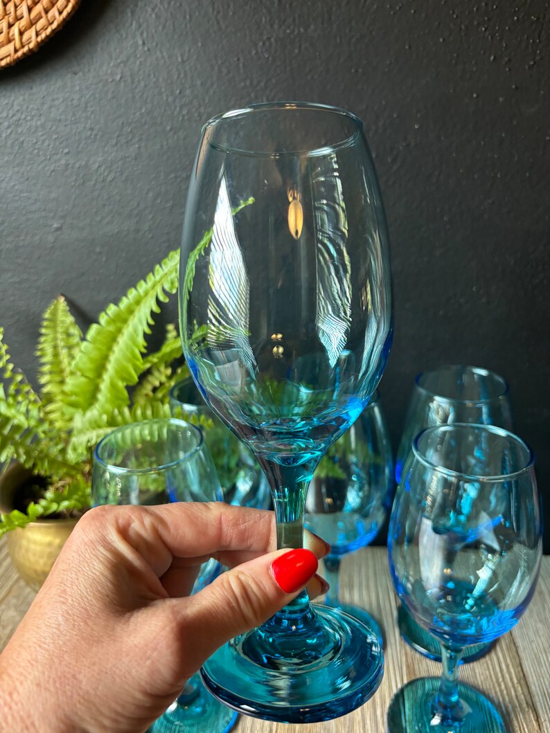 May include: Set of six blue glass wine glasses. The glasses have a clear, transparent body and a blue base. The glasses are arranged on a wooden surface.