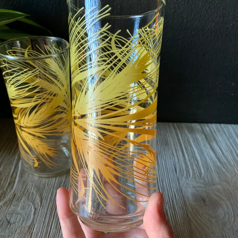 May include: Two clear glass drinking glasses with a yellow palm tree design. The glasses are different sizes.