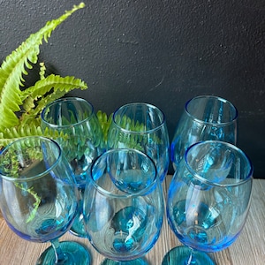 May include: Six blue glass wine glasses with a teal tint. The glasses are arranged in a circle on a wooden surface.